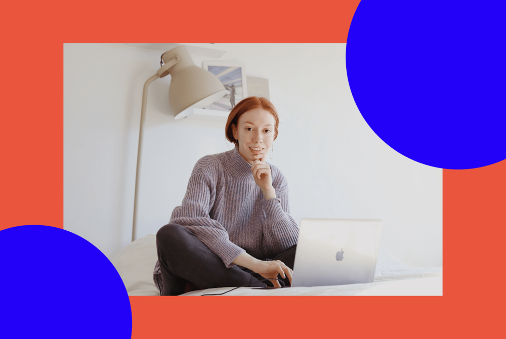 A woman working from home is sitting on a bed using a laptop computer.