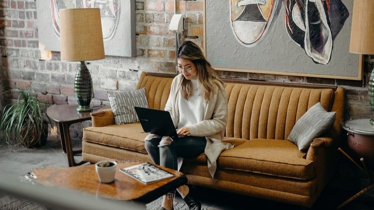 A woman in a seater sitting on a couch working remotely on her laptop.