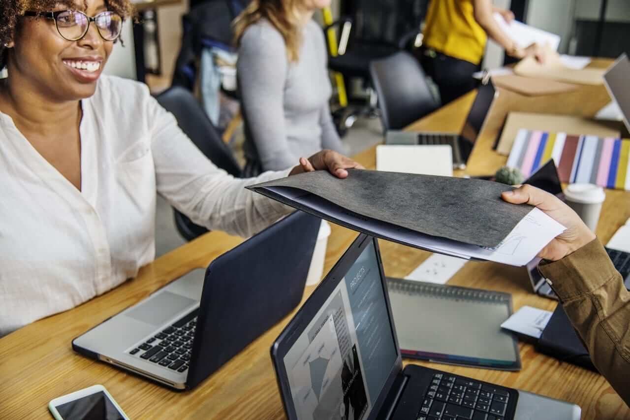 a woman handing a folder a man in front of a laptop computer.	