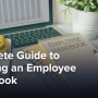 A laptop open on a desk, surrounded by notebooks, an employee handbook, coffee, and pencils